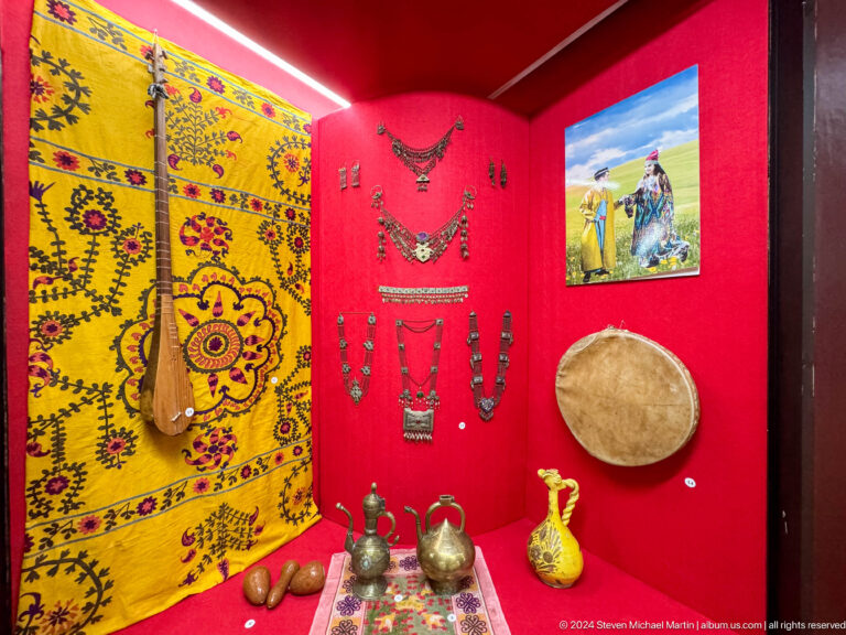 Medium view of a red-backed museum display showing traditional household and costume objects at the Central State Museum of Kazakhstan in Almaty.