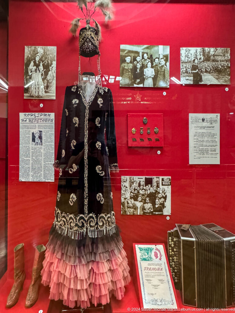 Medium view of a long dark historic embroidered coat displayed against a red backdrop in the Central State Museum of Kazakhstan in Almaty.