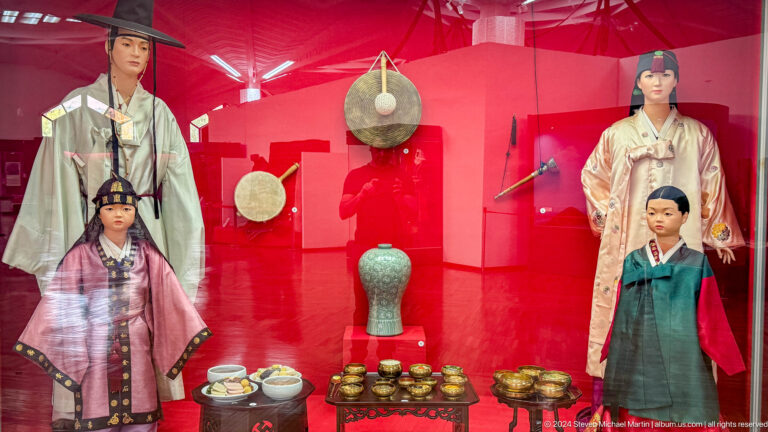 Medium view of a red museum display case with mannequins, vessels, a drum, and household objects in the Central State Museum, Almaty, Kazakhstan.