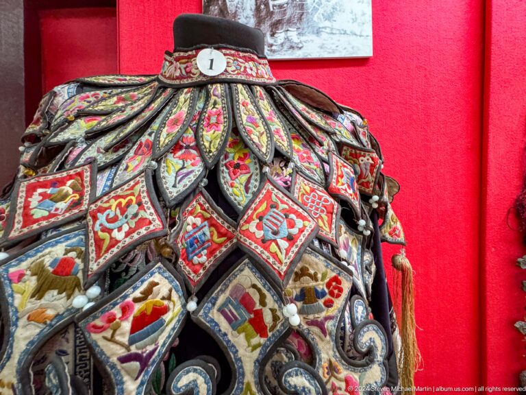 Close-up of a multicolored embroidered women’s bodice on red museum display backing, showing detailed panels and stitching.