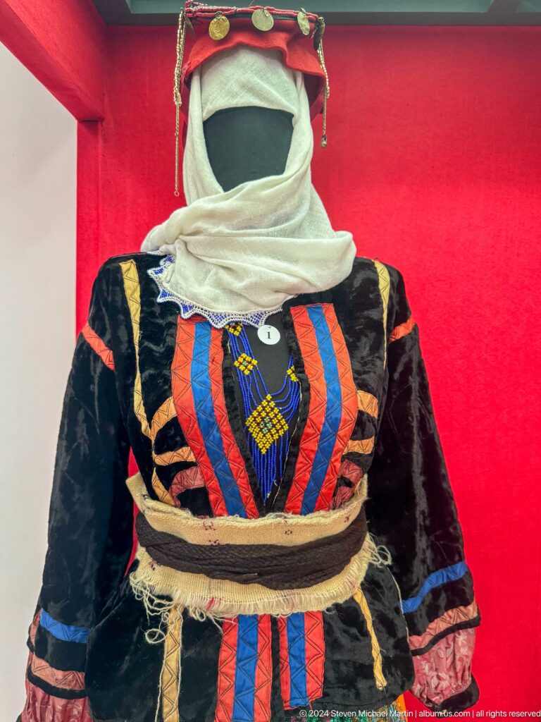 Colorfully paneled black traditional dress with bright vertical orange, blue, and red bands displayed against a red museum backing.
