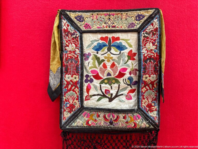 Medium view of a square embroidered textile panel with a central floral motif against a red museum display backing.