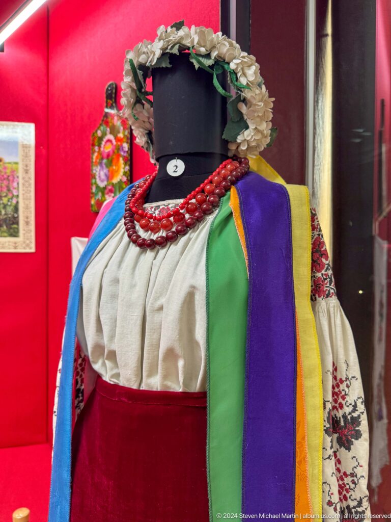 Women’s traditional costume with white blouse, red bead necklace, and multicolored shoulder drape displayed against a red background in the Central State Museum in Almaty, Kazakhstan.