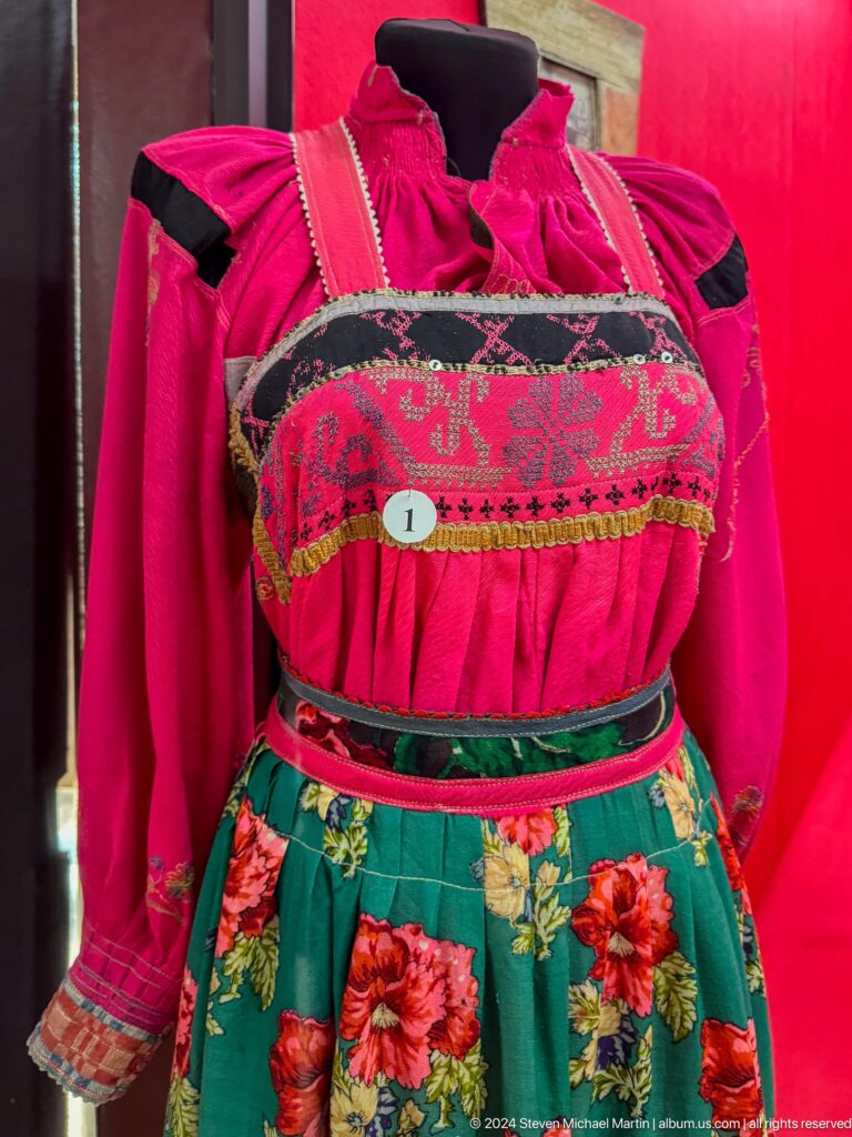 Medium view of a pink and green women’s embroidered dress with a densely decorated bodice displayed against a red backdrop in the Central State Museum in Almaty, Kazakhstan.