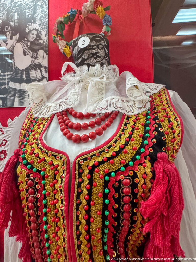 Women’s white embroidered blouse with a large red bead necklace on exhibit against a red museum display backing.