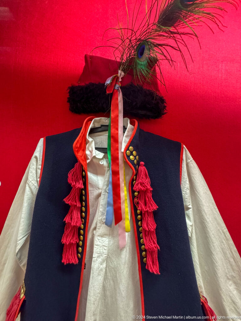 White traditional jacket with black, red, and yellow trim and tasseled front on display in a red exhibition case at the Central State Museum in Almaty, Kazakhstan.