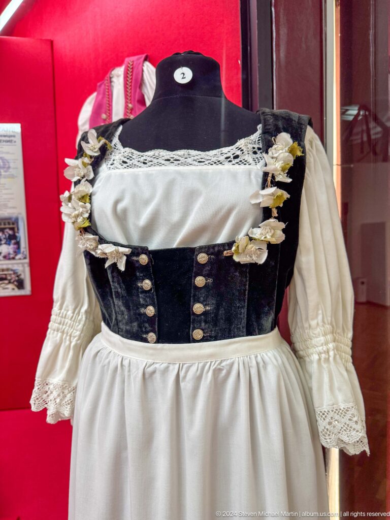 Museum display of a dark blue women’s bodice covered with coin-like ornaments over a long-sleeved white blouse on a mannequin.