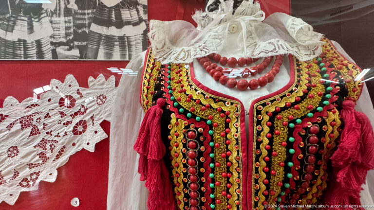 Close-up of a women’s costume showing an embroidered bodice over a white blouse with layered red bead necklaces against a red museum display background.