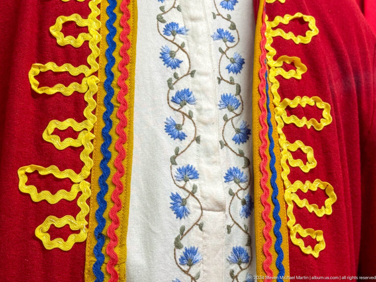 Close-up of blue floral embroidery on a white shirt beneath a red jacket displayed in a museum.