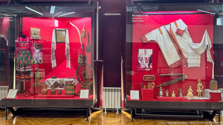 Medium view of a red museum display case holding white garments, weapons, vessels, and textiles in a regional ethnographic exhibition at the Central State Museum of Kazakhstan in Almaty.