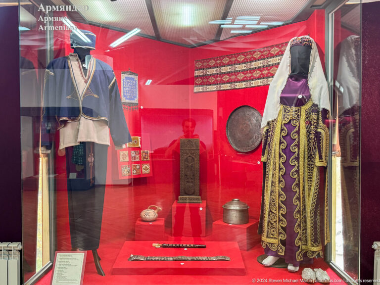 Red museum display case with a long embroidered robe, drum, and household objects at the Central State Museum of Kazakhstan in Almaty.