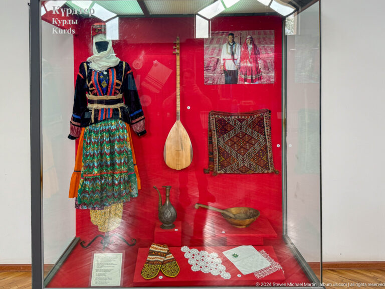 Museum display case with a colorful traditional dress, stringed lute, vessel, and woven bag against a red background.