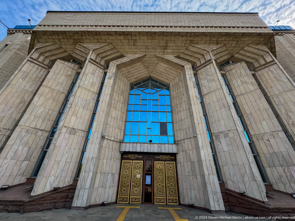 Low-angle view of the modern main entrance of the Central State Museum in Almaty, Kazakhstan, showing a recessed doorway beneath blue windows.