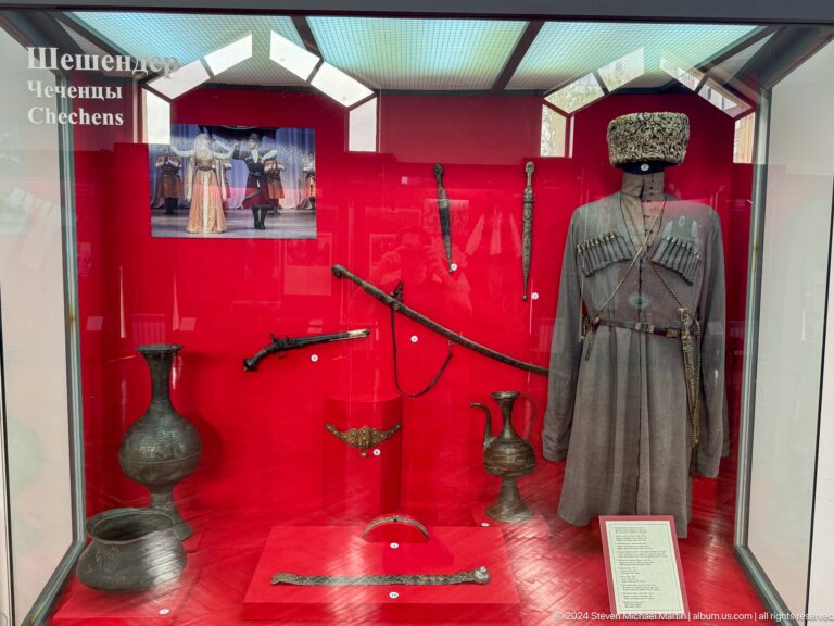Historic coat, firearms, headwear, and metal vessels displayed in a red case at the Central State Museum in Almaty, Kazakhstan.