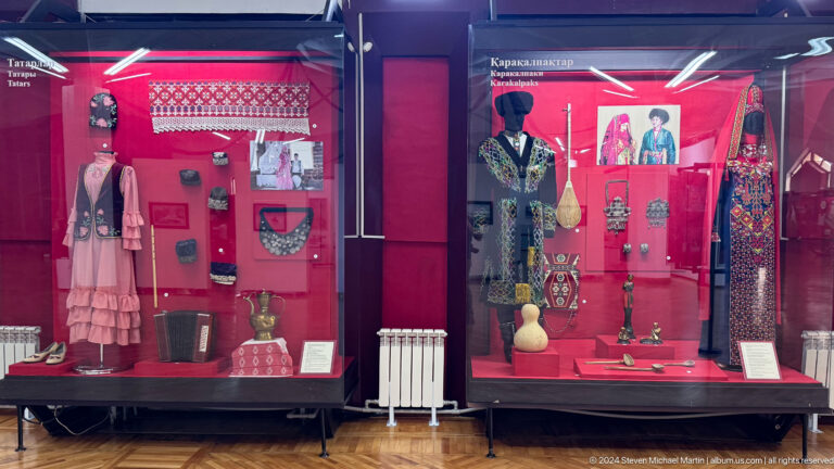 Two red museum display cases holding traditional regional clothing, jewelry, and framed portraits in a gallery at the Central State Museum of Kazakhstan.