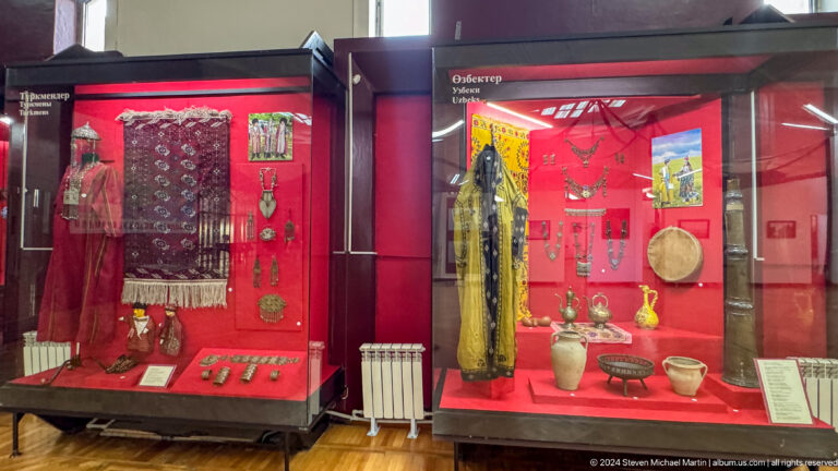 Red museum display cases with regional textiles, a long robe, and domestic vessels in the Central State Museum in Almaty, Kazakhstan.