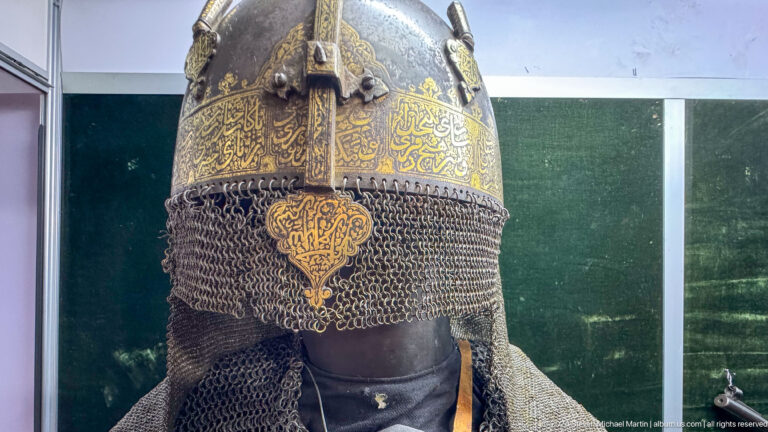 Ornate gold-colored ceremonial helmet with protective metal elements displayed against a red backdrop in the Central State Museum of Kazakhstan in Almaty.