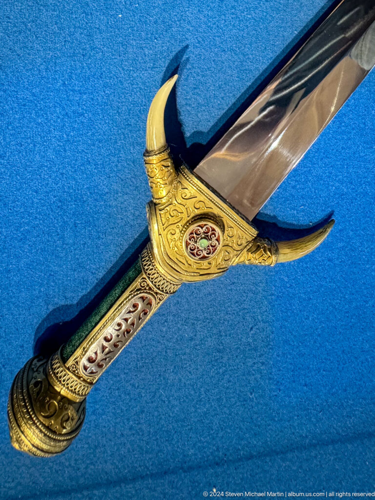Close-up of a curved ornate saber hilt and matching scabbard decorated with gold-colored metalwork and fine patterns in a museum display.
