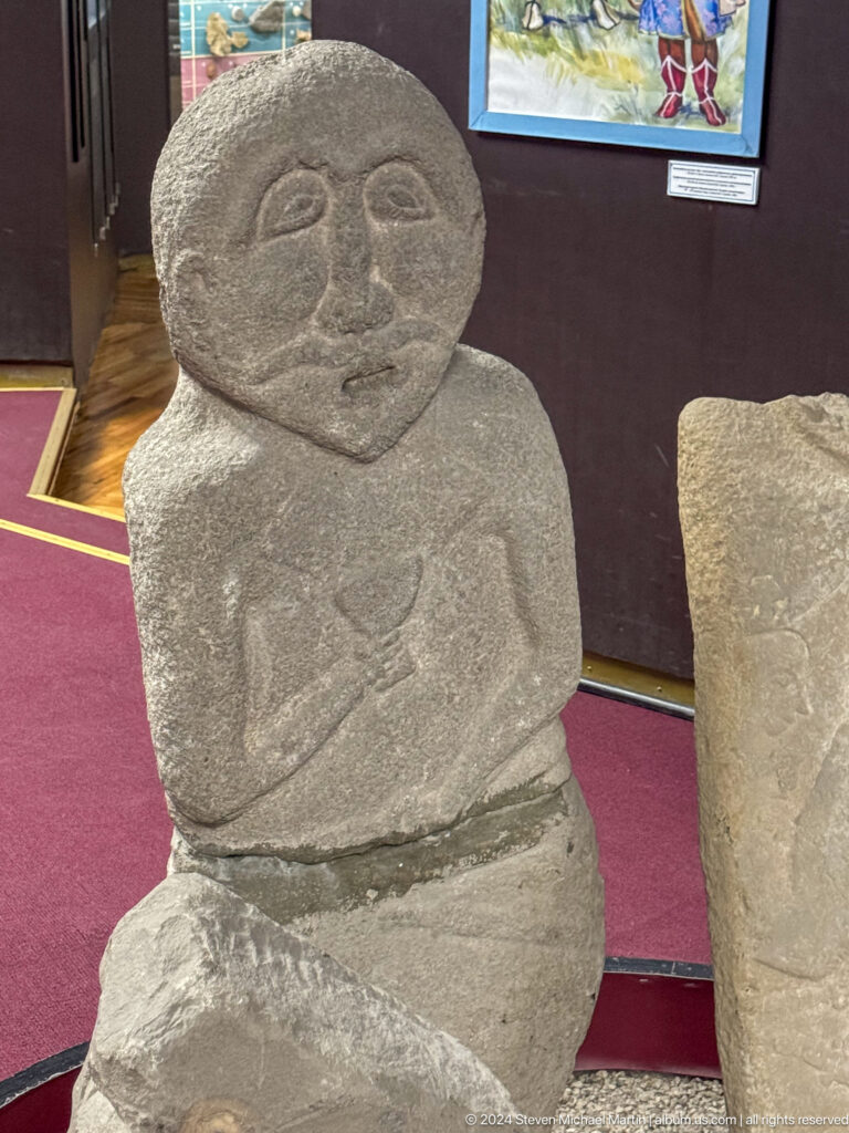 Medium view of a small gray anthropomorphic stone figure with folded arms displayed on red fabric in a museum setting.