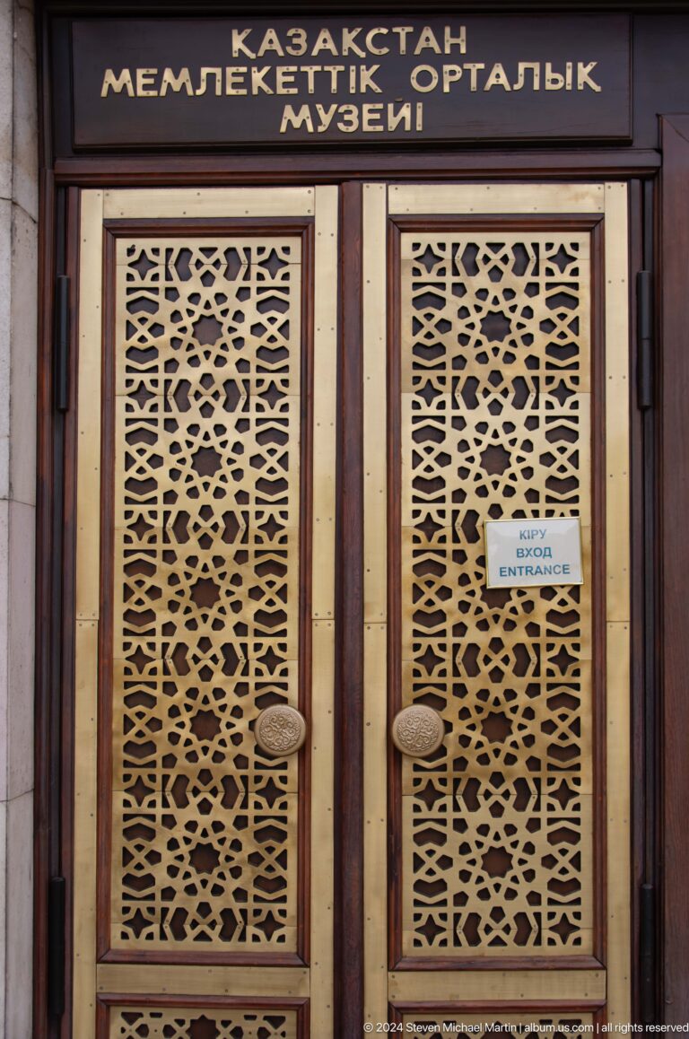 Ornate geometric metal entrance doors with a small bilingual sign at the Central State Museum of Kazakhstan in Almaty.