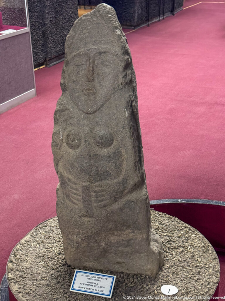 Medium view of an upright anthropomorphic stone stele with a carved human face and torso at the Central State Museum in Almaty, Kazakhstan.