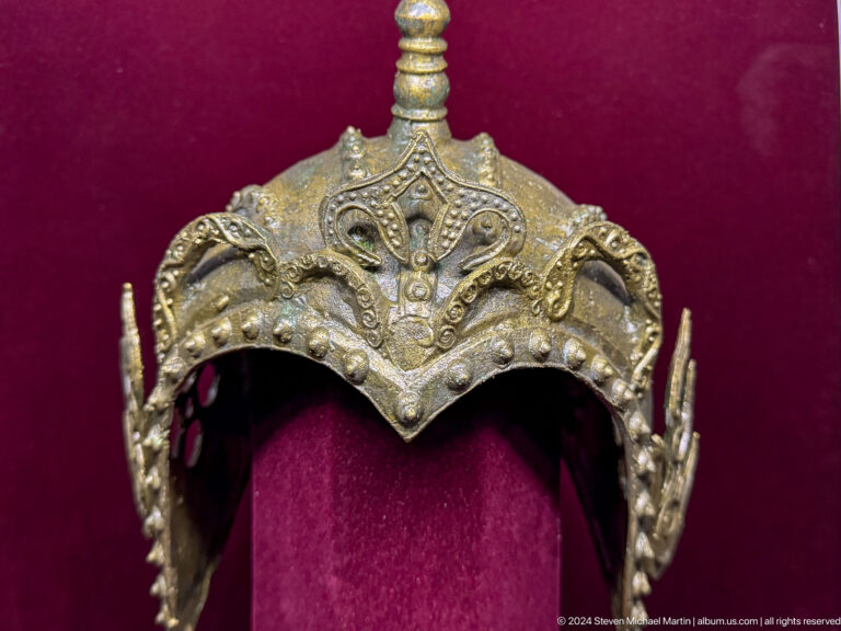 Ornate gold-colored ceremonial metal headdress displayed against red backing at the Central State Museum in Almaty, Kazakhstan.