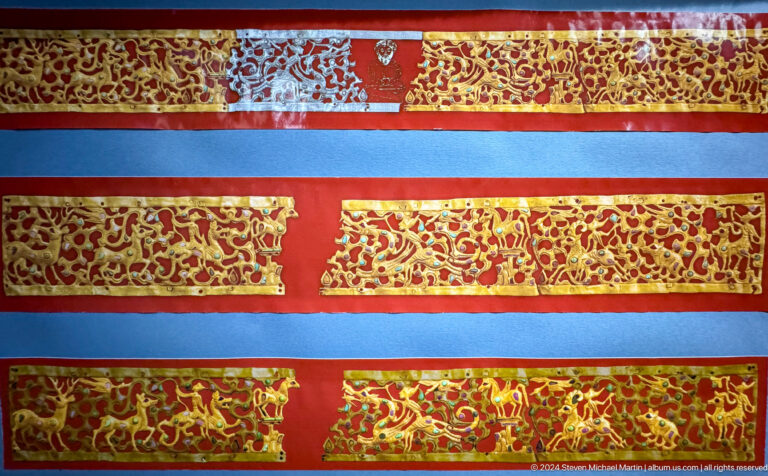 Medium view of red and blue textile bands densely embroidered with gold on a red museum display backing.