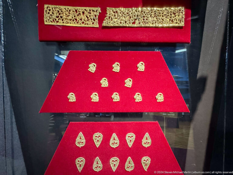 Medium view of small gold ornaments and appliques arranged on red fabric in a display case at the Central State Museum of Kazakhstan in Almaty.