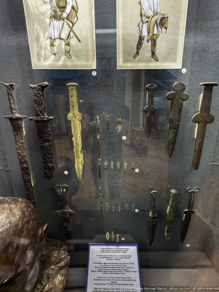 Medium view of a museum display case showing historic swords, daggers, and other small metal weapons at the Central State Museum of Kazakhstan in Almaty.