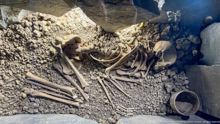 Museum display of an excavated burial with a human skeleton laid out in a pit, accompanied by pottery vessels and other grave goods.