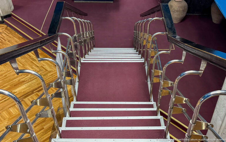 Curved red-carpeted staircase with metal railings inside the Central State Museum of Kazakhstan, with exhibition walls and display cases visible.