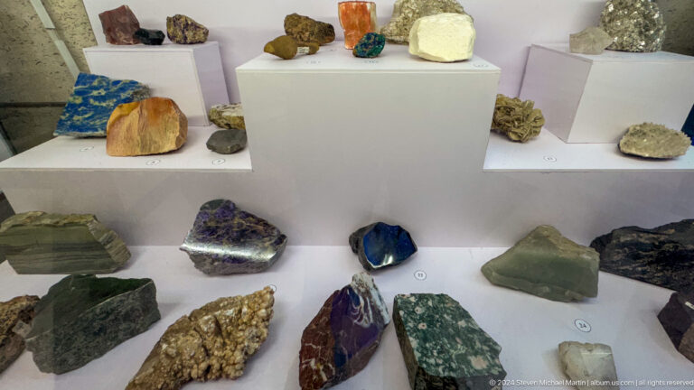 Medium view of polished and rough mineral specimens arranged on white shelves in a museum display at the Central State Museum of Kazakhstan in Almaty.