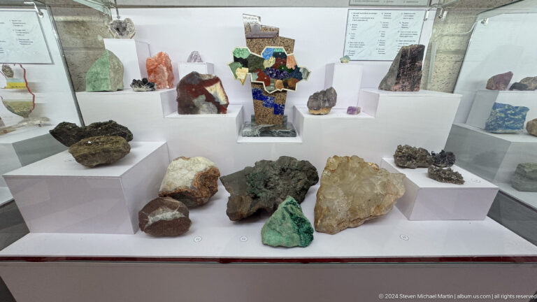 Medium view of multicolored mineral specimens on tiered white plinths against a red background in a geology display at the Central State Museum in Almaty, Kazakhstan.