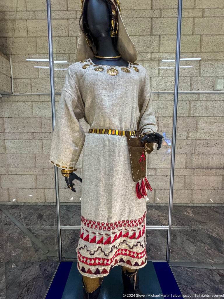 Mannequin wearing a plain light-colored traditional Central Asian male tunic with belt and side pouch in a museum display.