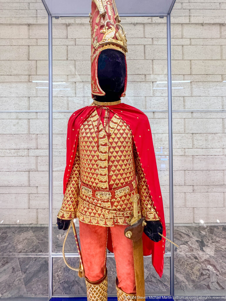 Mannequin wearing a red traditional female garment with prominent gold detailing, displayed against a red backdrop in the Central State Museum of Kazakhstan in Almaty.
