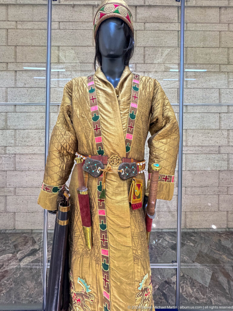 Mannequin wearing a tan quilted traditional coat with colorful embroidered trim, displayed against a red background in the Central State Museum in Almaty, Kazakhstan.
