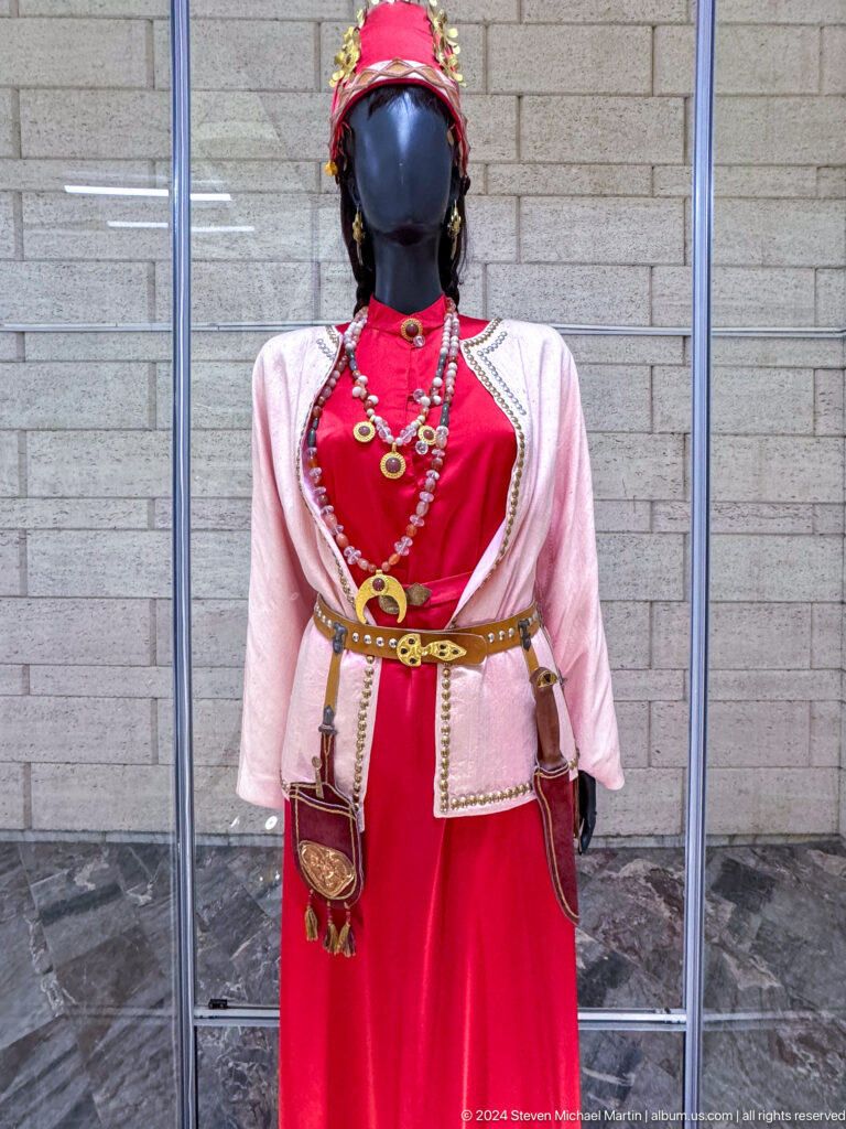 Traditional women’s costume on a mannequin, featuring a white blouse, red vest, and jewelry, displayed in a museum case with a red background.