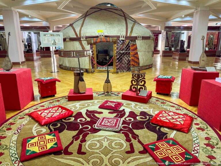 Circular museum display of Kazakh yurt furnishings with patterned textiles and household items arranged to suggest a nomadic interior.
