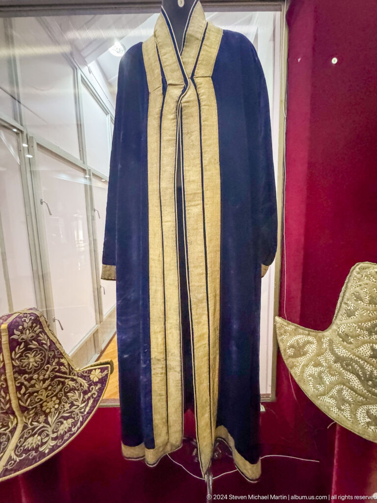 Dark ceremonial robe with wide vertical gold trim displayed on a hanger against a plain museum backdrop.