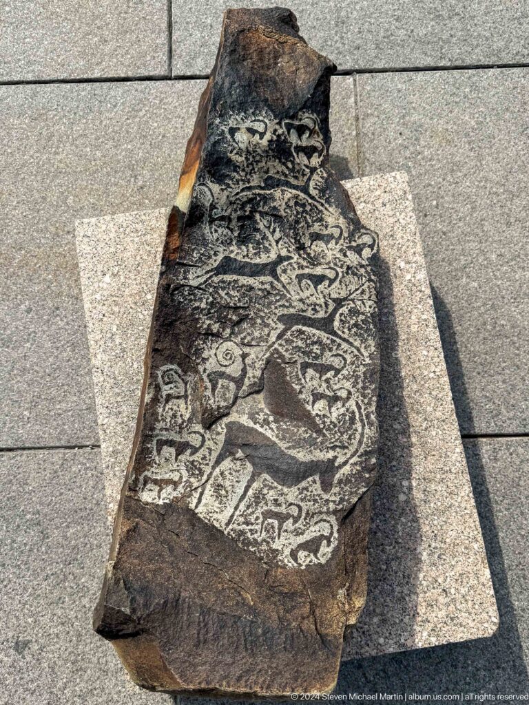 Dark carved stone slab with petroglyph-style animal and human-like figures incised on its rough surface.
