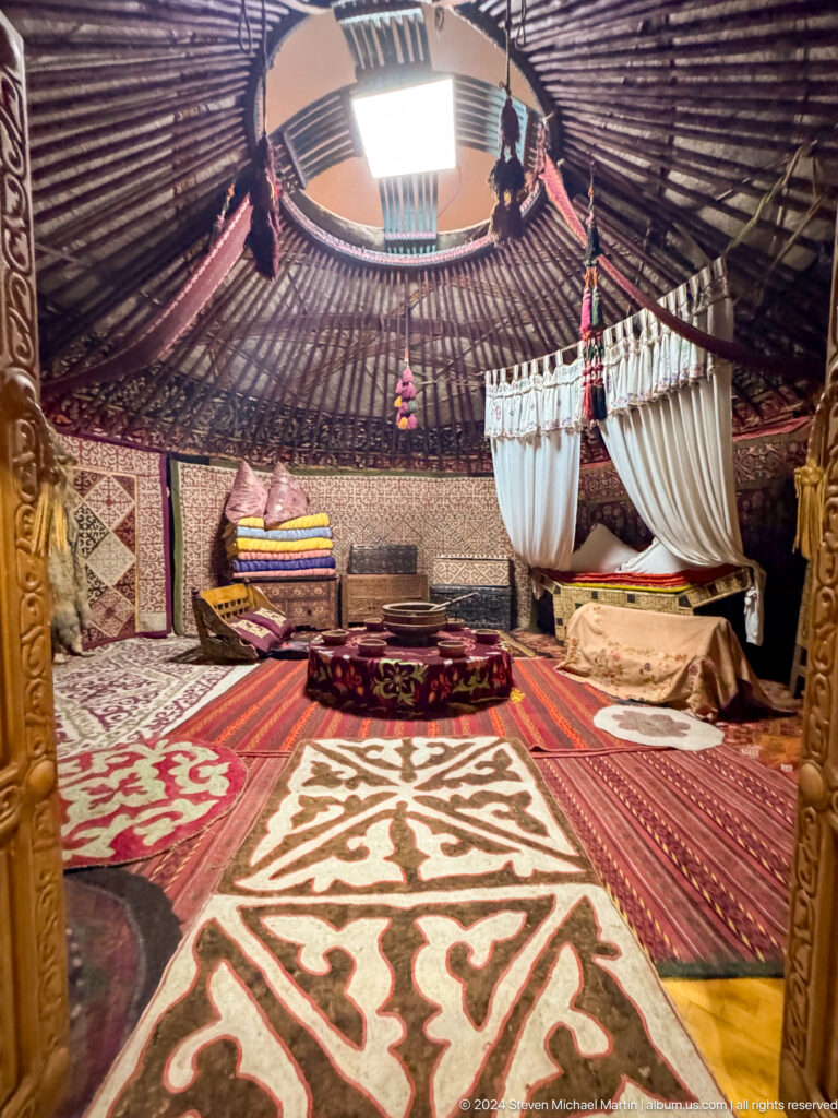 Medium view of a furnished traditional Kazakh yurt interior with textiles, cradle, and low table in a museum exhibit.