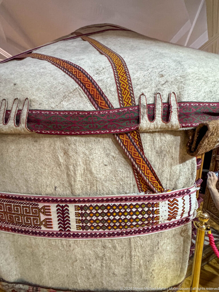 Close-up of a felt yurt wall with red woven straps crossing over a wooden lattice frame against a red exhibition backing.