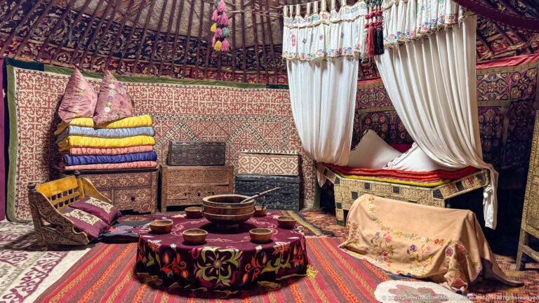 Ornate Kazakh yurt interior in a museum display with carpets, patterned textiles, and a curtained sleeping area under a circular roof.