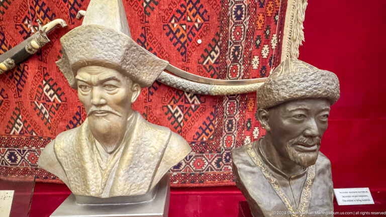 Two sculpted male portrait busts wearing different traditional hats displayed together in a museum setting.