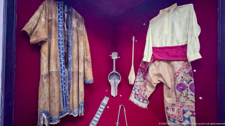 Medium view of a museum display showing a traditional clothing ensemble of shirts, trousers, boots, and a small vessel on a red backing at the Central State Museum in Almaty, Kazakhstan.