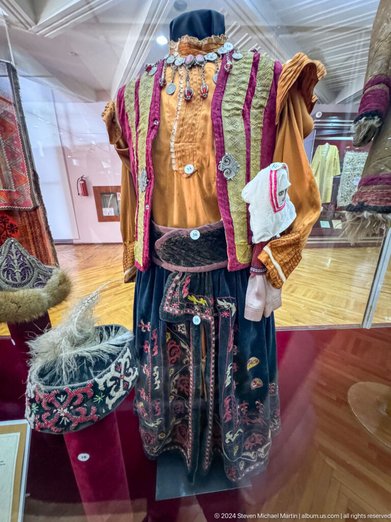 Traditional Kazakh women’s winter outfit with fur-trimmed garments, embroidered fabrics, and a small bag displayed on a museum mannequin.