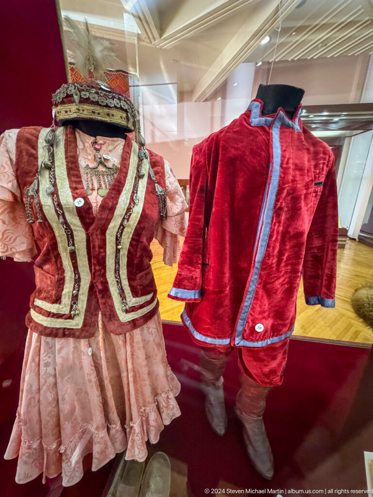 Two traditional velvet jackets, one red and one brown with light-colored trim, displayed on hangers against a red museum wall.