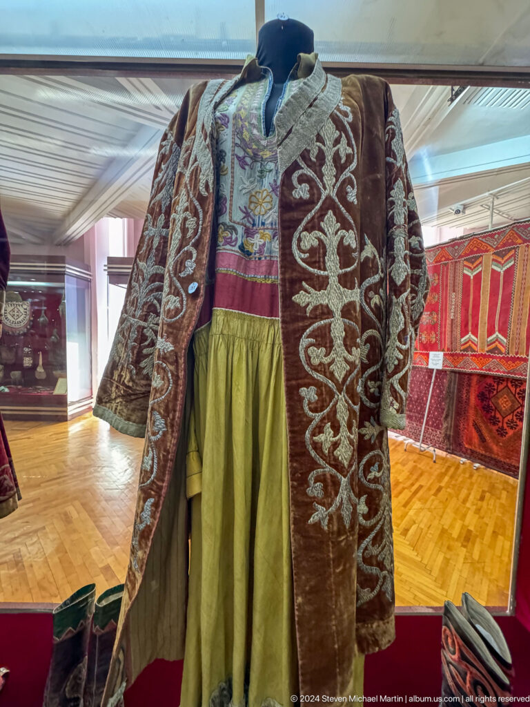 Embroidered dark ceremonial robe with white scrolling patterns displayed over a yellow dress on a mannequin in a museum setting.