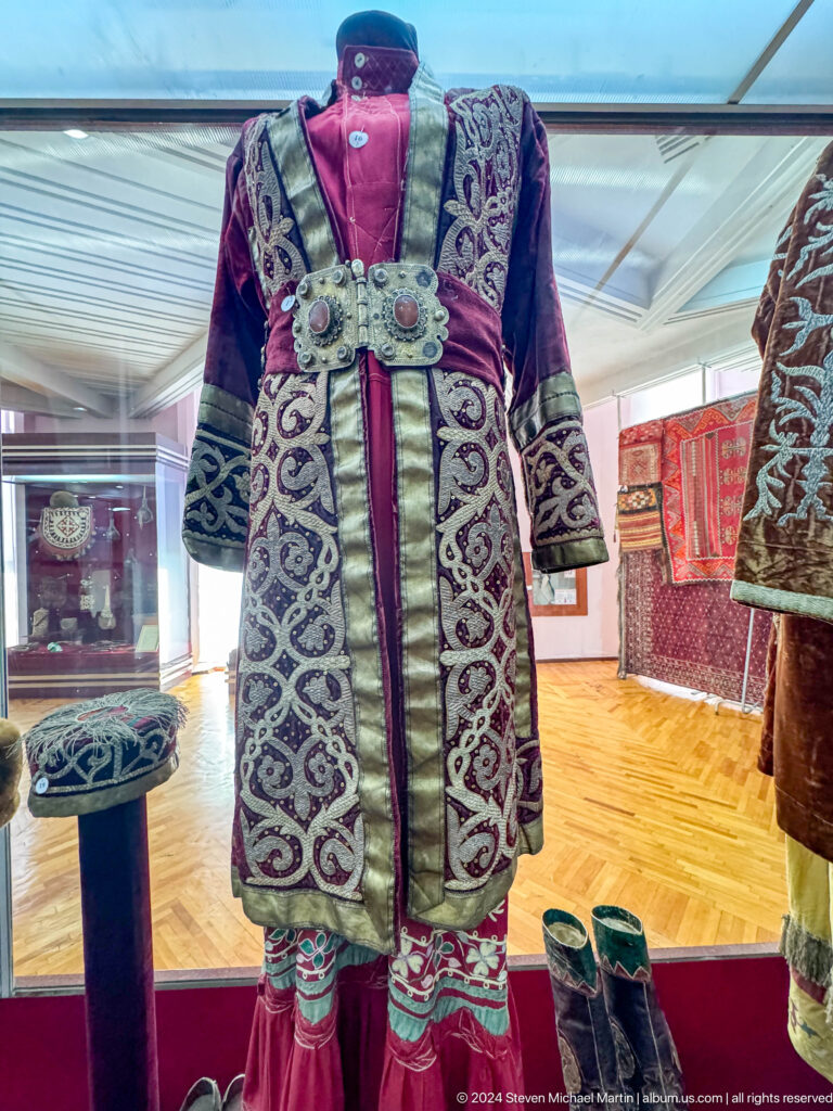Blue and white embroidered traditional coat with dense curving patterns displayed upright in a museum case.