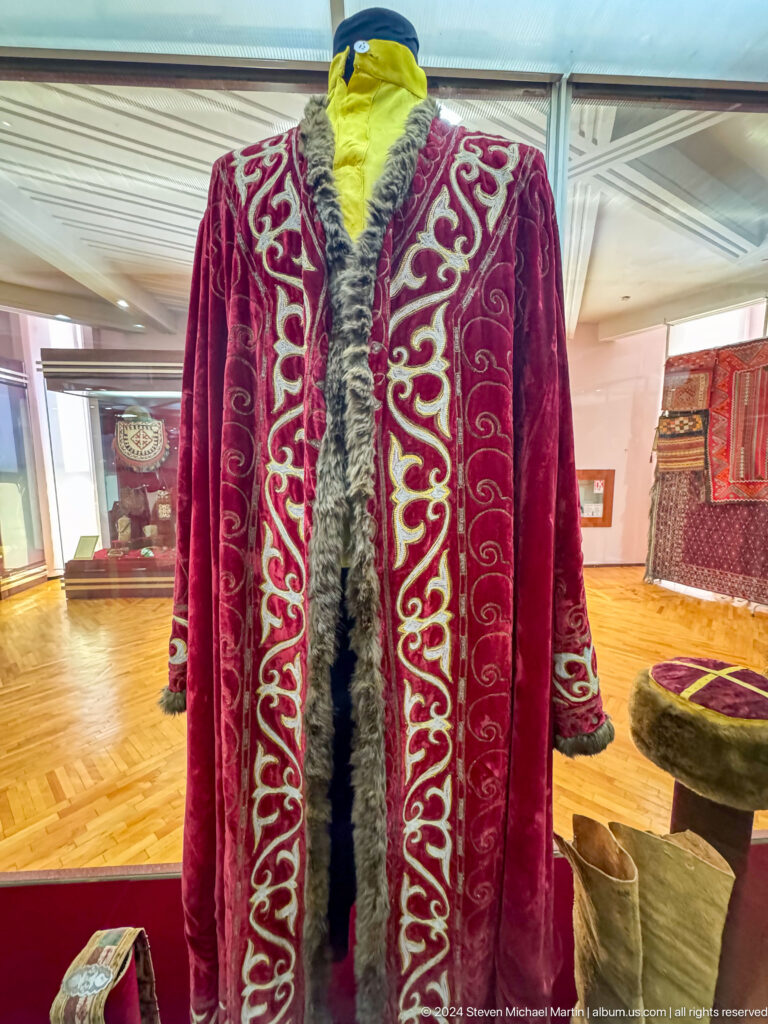 Close-up of red velvet garment with dense white scroll embroidery displayed against a red museum backing.